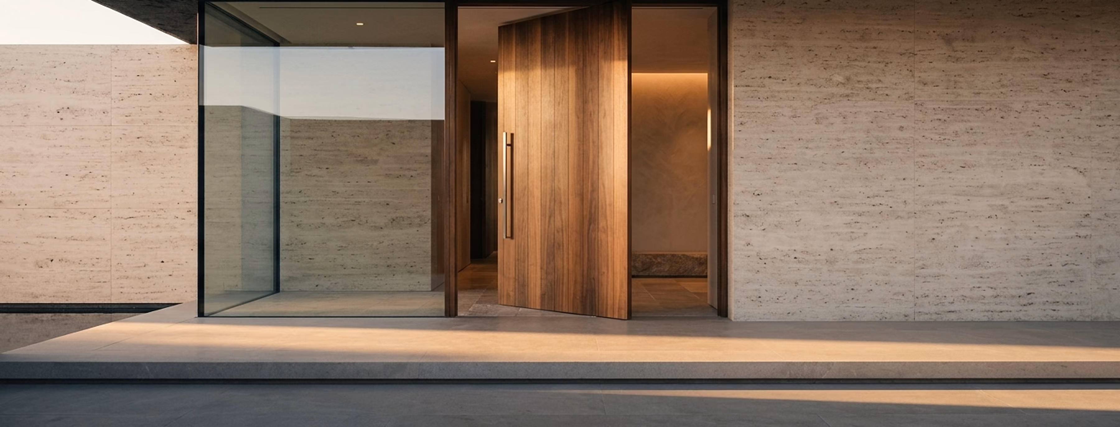 The entrance of a luxury villa at sunrise: a tall pivoting walnut door standing slightly open onto a warm interior.