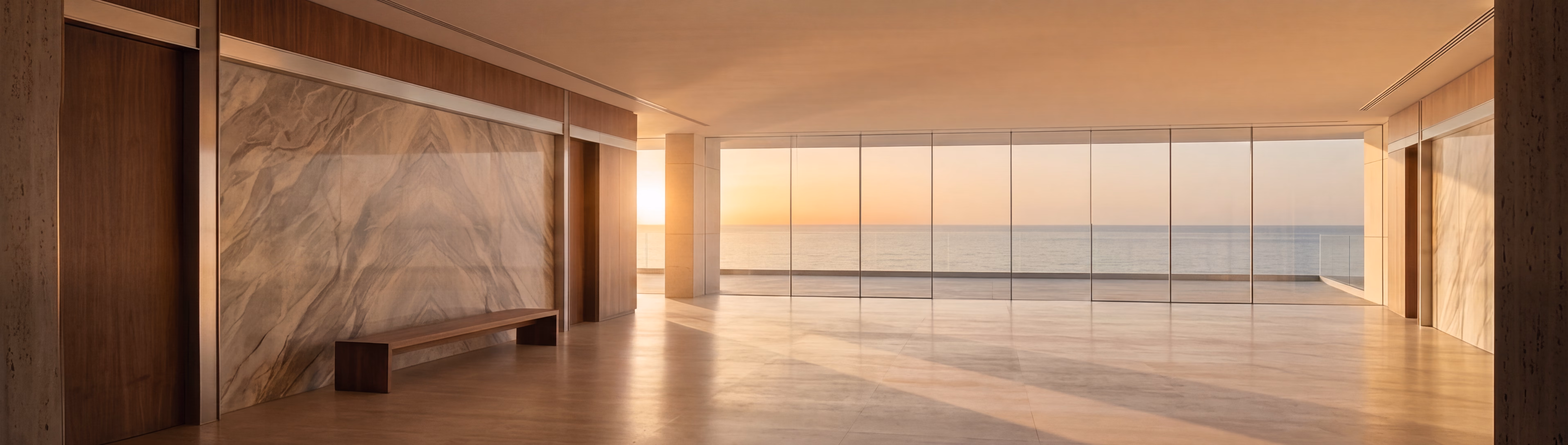 An empty sunlit interior at sunrise, a book-matched marble wall and timber bench along the near side, the full-height glazed wall opening onto the sea beyond.